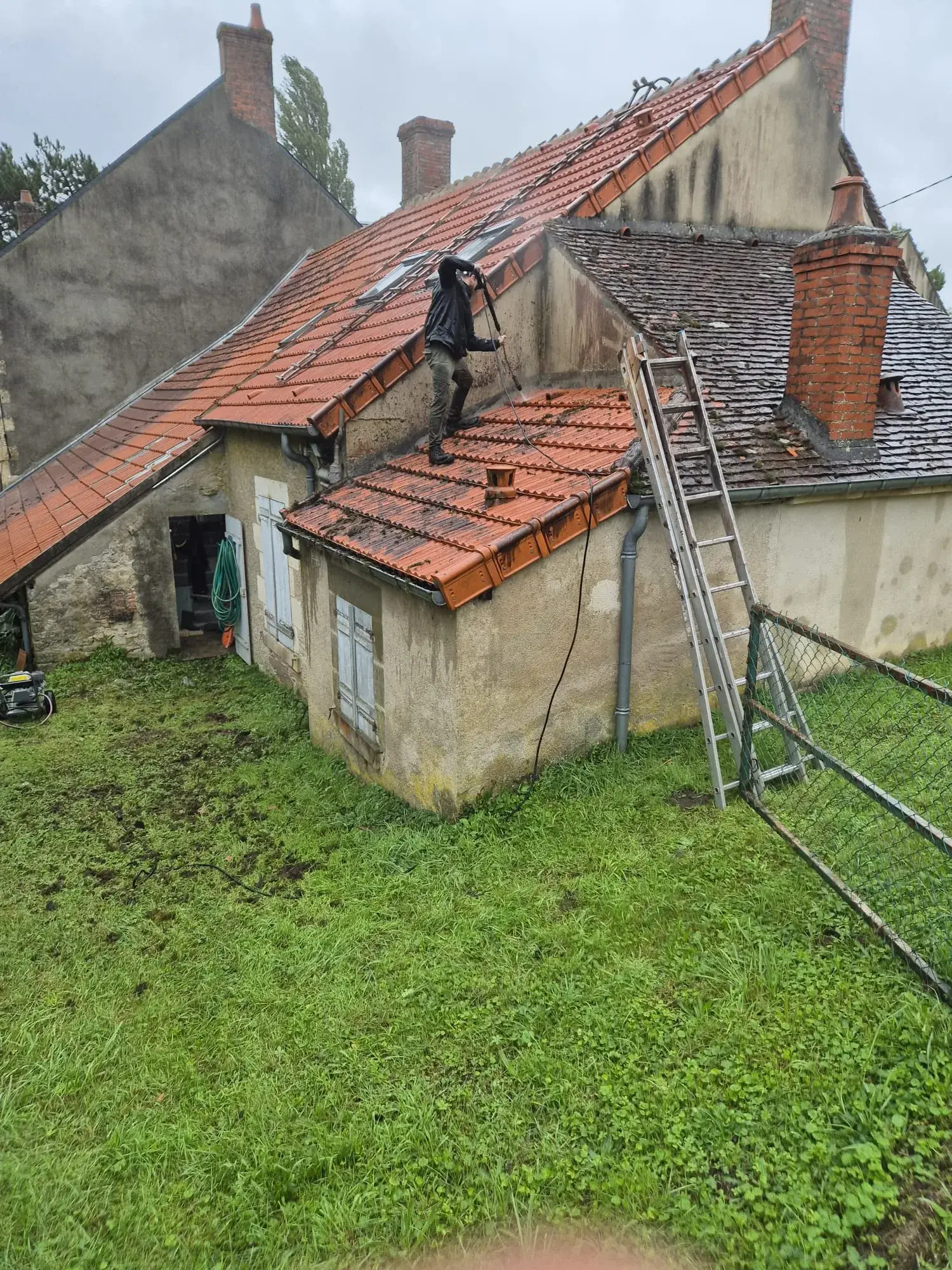 Couvreur nettoyant une toiture en tuiles mécaniques au nettoyeur haute pression, pan voisin encore couvert de mousse